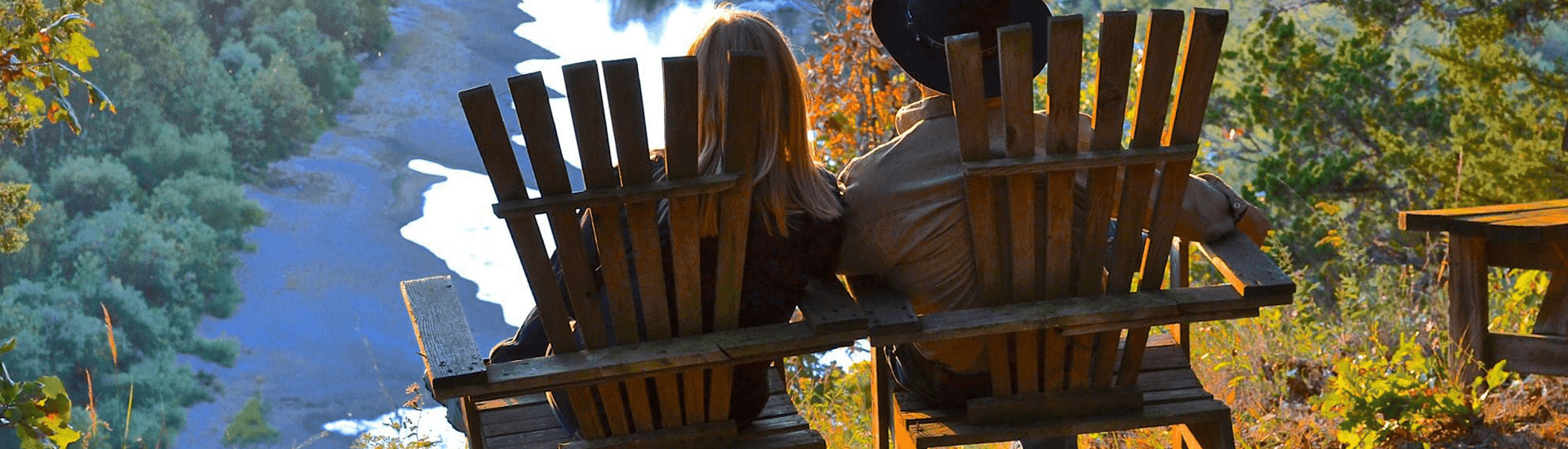 A couple sits side by side in wooden chairs, overlooking a serene river surrounded by trees.