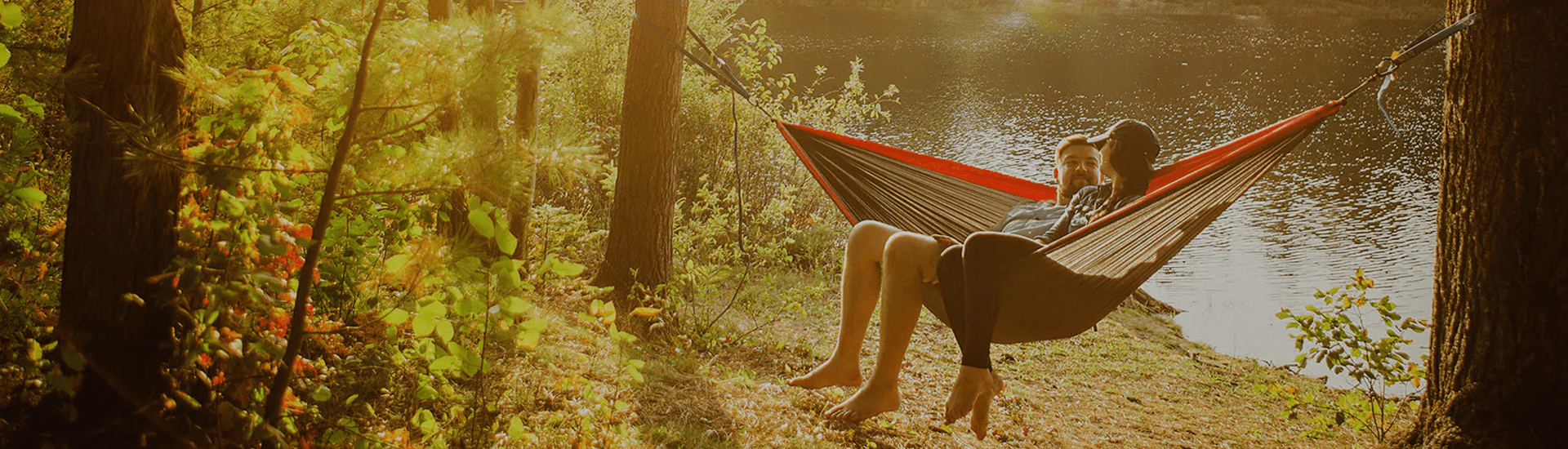 A couple relaxing in a hammock by a tranquil lake surrounded by trees.