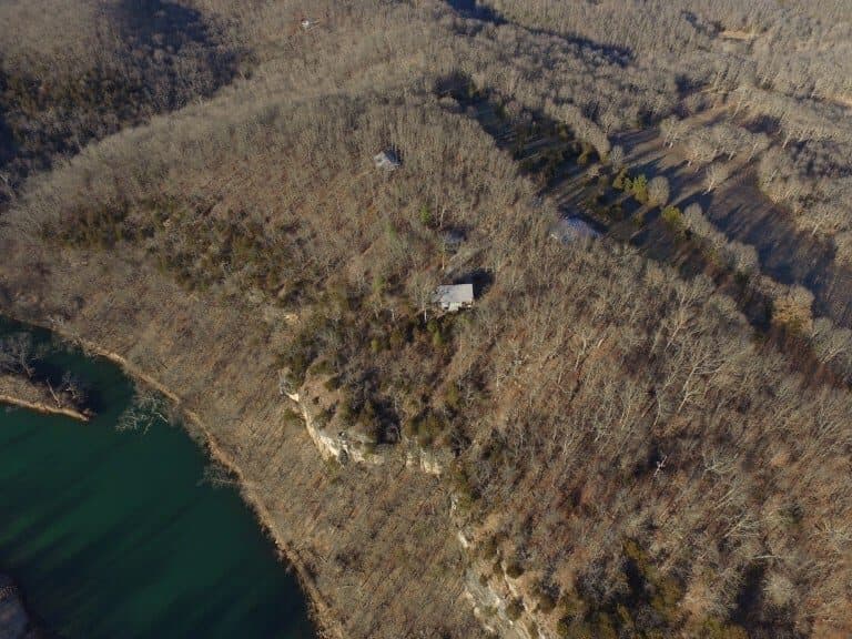 Aerial view of a wooded area with a river, showcasing a sparse landscape and a single house located on a hilltop.