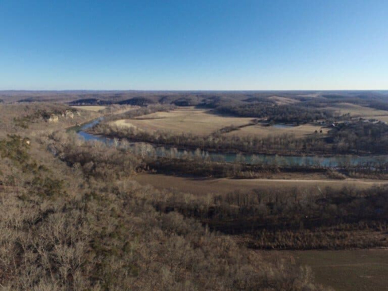 Aerial view of a winding river surrounded by bare trees and rolling hills under a clear blue sky.